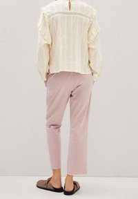 Cremfarbene Bluse mit strukturiertem Stoff und Rüschensäumen, Rückenschlitzdetail, kombiniert mit hellrosa Hosen und braunen Slippern.