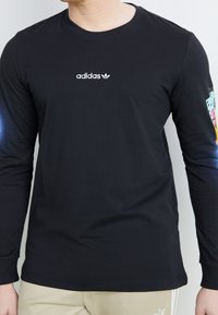 Czarna koszulka Adidas z długim rękawem i białym logo na piersi, z kolorową grafiką na prawym rękawie, noszona przez osobę w beżowych spodniach.