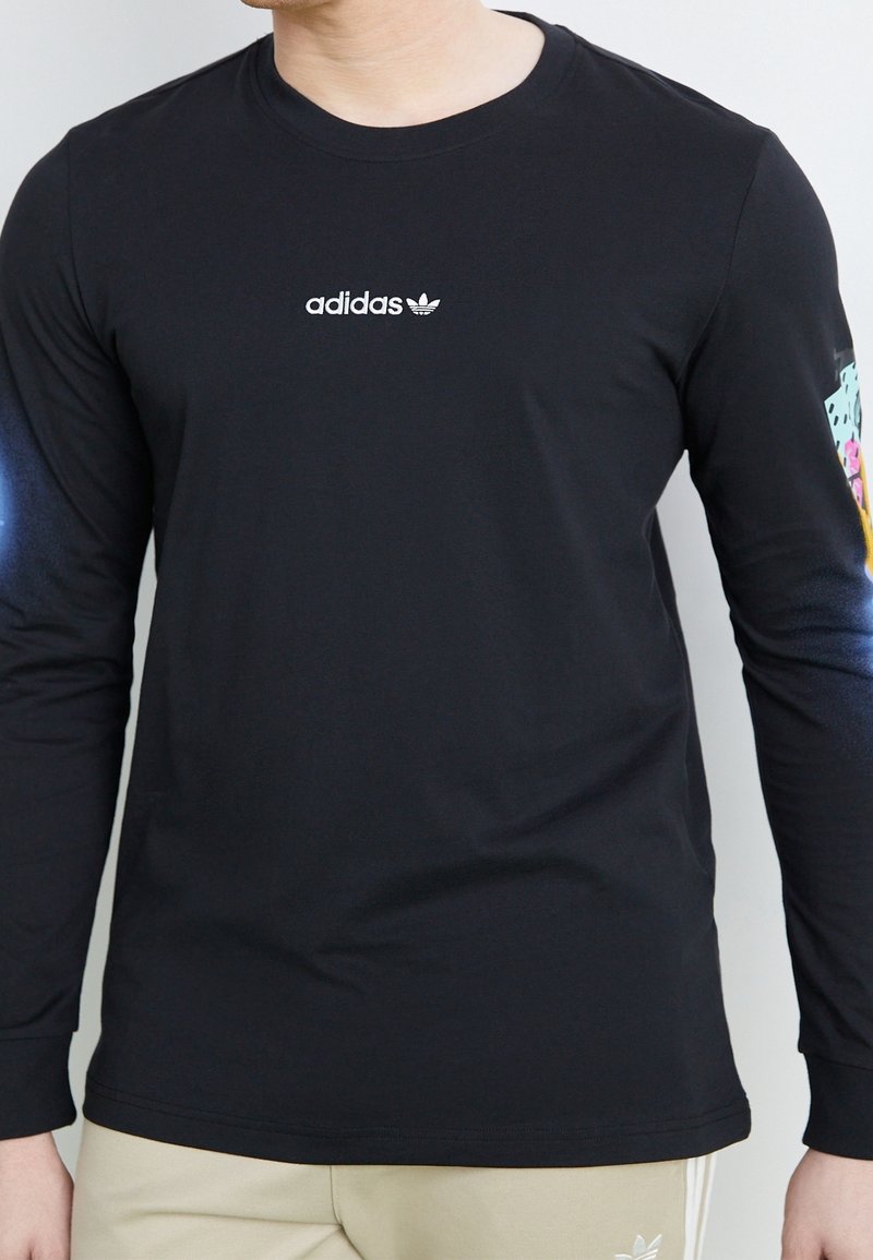 Czarna koszulka Adidas z długim rękawem i białym logo na piersi, z kolorową grafiką na prawym rękawie, noszona przez osobę w beżowych spodniach.