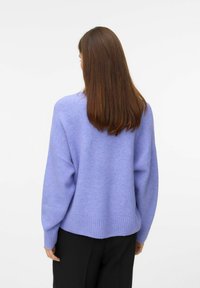 Oversized sweter w kolorze lawendowym z prążkowanym wykończeniem, długimi rękawami i luźnym krojem. Miękka tekstura, noszony na ciemnych spodniach.