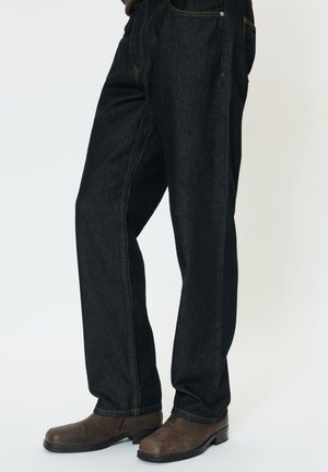 Personne portant un jean droit bleu foncé et des bottines en cuir marron, debout sur un fond clair.