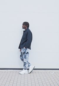 Fekete, oversized dzseki gallérral; világosszürke nadrág sötét absztrakt mintával; fehér, vastag talpú sportcipők; világos háttér előtt áll.