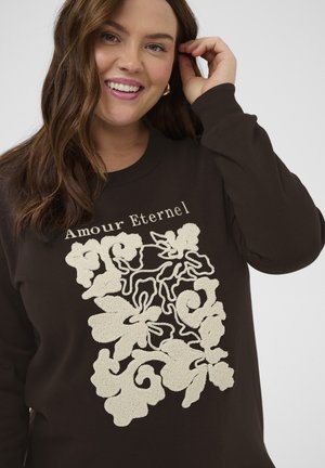 Smilende kvinde med bølget brunt hår iført mørk sweatshirt med cremefarvet blomsterbroderi og teksten "Amour Eternel" på brystet.