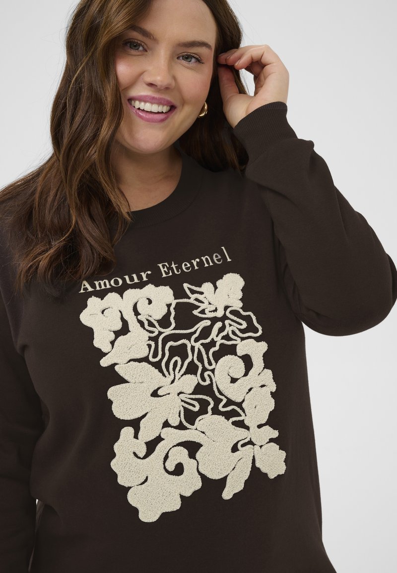 Femme souriante aux cheveux bruns ondulés porte un sweat-shirt foncé orné d’une broderie florale crème et du texte « Amour Eternel » sur la poitrine.