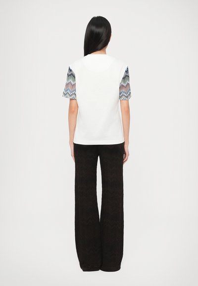 Λευκή μπλούζα με κοντά μανίκια σε σχέδιο μπλε, πράσινου και ροδακινί. Μαύρο παντελόνι με φαρδιά μπατζάκια και υφασματικό ζιγκ-ζαγκ μοτίβο.