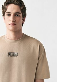 Camiseta de algodón beige con cuello redondo y mangas cortas, con impresión negra que dice "AMSTERDAM DE NEGEN STRAATJES" en la parte frontal.