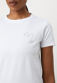 Maglietta bianca in cotone con un piccolo motivo di strass luccicanti sul lato sinistro del petto, scollo a girocollo e maniche corte.