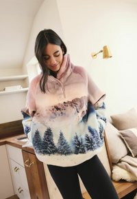 Fleece pullover in gradient pink to blue, featuring a mountain and forest print, with a high collar and soft texture. Black leggings complete the look.