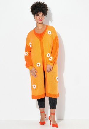 Langoranje cardigan gebreid met bloemenapplicaties, met een V-hals, knopen en zakken. Gecombineerd met zwart gebloemde leggings en oranje hakken.