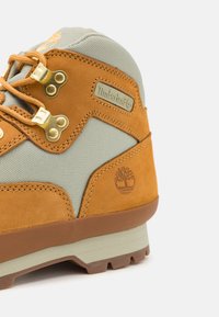 Bota de senderismo marrón y gris con parte superior de cuero, detalles de tela, herrajes dorados y suela de goma con patrón de tacos. Logo de Timberland exhibido.