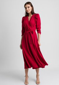 Czerwona sukienka o kroju kopertowym z rękawami o długości 3/4 i bufkami, z wiązanym w pasie biodrem oraz zwiewnym dołem. Gładki, lekki materiał podkreśla opadanie tkaniny.