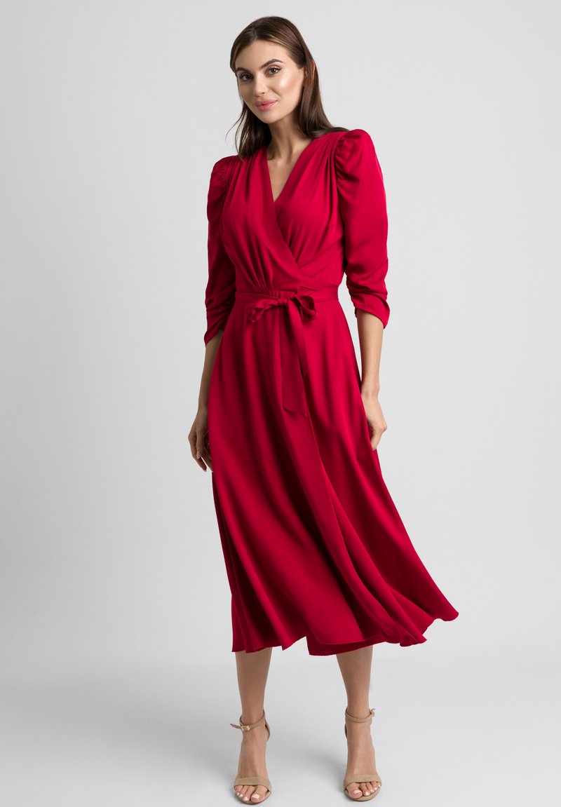 Czerwona sukienka o kroju kopertowym z rękawami o długości 3/4 i bufkami, z wiązanym w pasie biodrem oraz zwiewnym dołem. Gładki, lekki materiał podkreśla opadanie tkaniny.