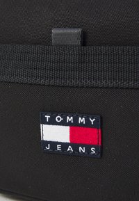 Black fabric bag with a woven texture; features a rectangular embroidered Tommy Jeans logo in blue, white, and red.