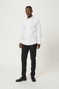 Cămașă albă cu mânecă lungă și închidere cu nasturi, din material simplu, cu guler ascuțit, purtată cu pantaloni negri și pantofi din piele. Fără modele sau broderii.