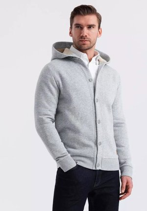 Sudadera gris hecha de tela suave, con una parte delantera abotonada, dos bolsillos y un forro interior beige visible en la capucha.