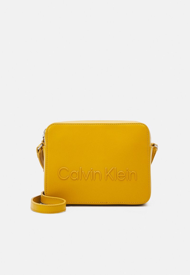 Gul læder crossbody-taske med rektangulær form, prægede "Calvin Klein"-logo, justerbar rem og glat tekstur.