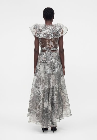 Γυναίκα που φοράει διαφανές φόρεμα μέγιστου μήκους με λουλουδάτα σχέδια, στρώσεις βολάν στους ώμους και ζώνη στη μέση, φωτογραφημένη από πίσω μπροστά σε απλό φόντο.