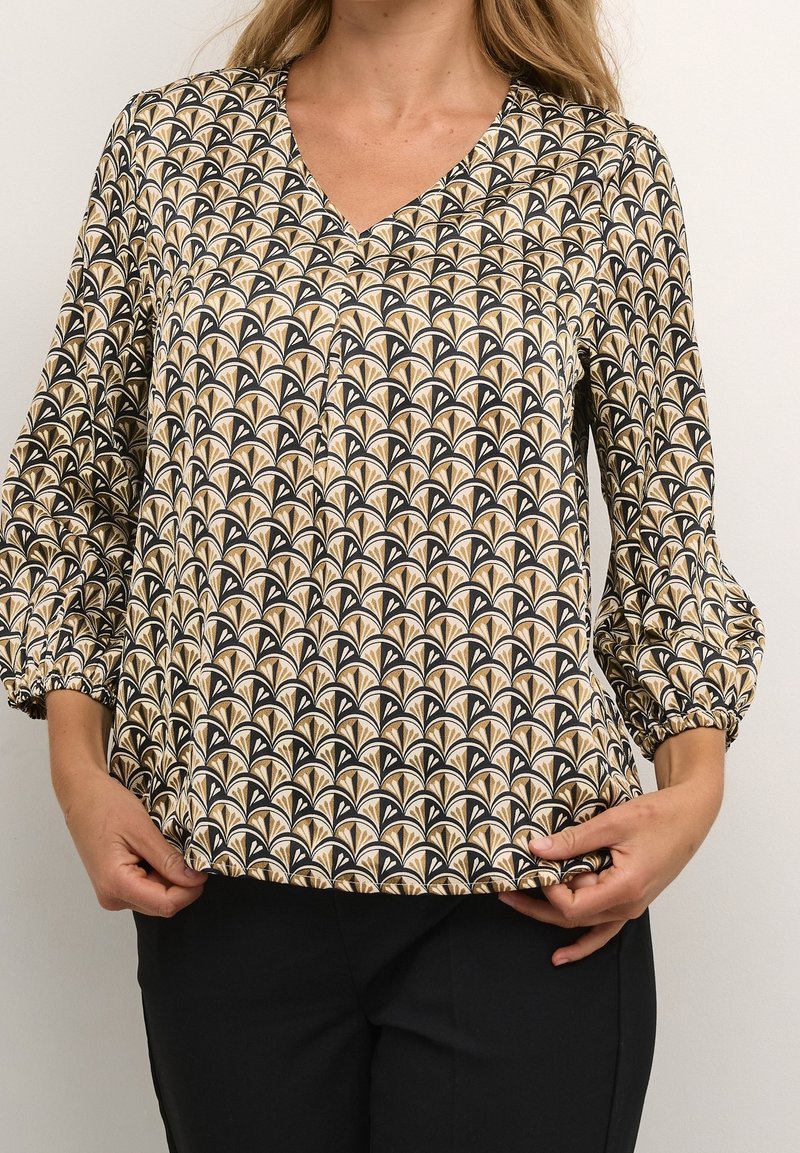 Mujer vestida con una blusa de manga larga de patrón beige, negro y blanco con puños abullonados y pantalones negros, sosteniendo el dobladillo de la blusa.