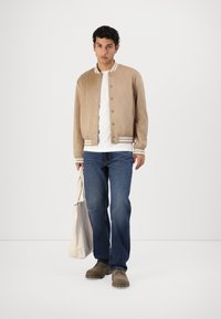 Lys beige bomberjakke med hvite stripete mansjetter, brukt over en hvit t-skjorte, kombinert med blå jeans og brune sko, mens han/hun bærer en beige tote bag.