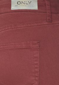 Tecido de denim vermelho com costura visível e um bolso. Apresenta uma etiqueta com a inscrição "ONLY" na cintura, indicando a marca e o estilo.