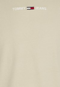 Světle béžové tričko s krátkým rukávem z bavlny, které má vyšívané logo "TOMMY JEANS" v bílé, červené a námořnické modré barvě.