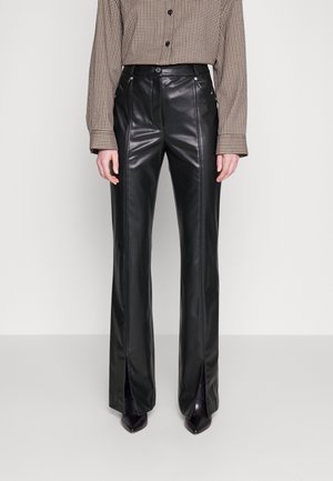 Vrouw draagt een zwarte faux-lederen broek met hoge taille, met voornaden en voorentrappijen over zwarte puntige schoenen, gecombineerd met een geruit bloesje met lange mouwen.