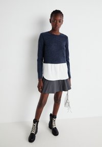Un pull marine avec un ourlet superposé blanc, une jupe grise évasée et des bottes noirs à lacets avec des œillets dorés. Le mannequin tient un sac à main argenté.