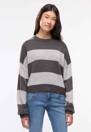 Donna con capelli lunghi e scuri che indossa un maglione a righe grigio e grigio chiaro e jeans blu, in piedi davanti a uno sfondo semplice.