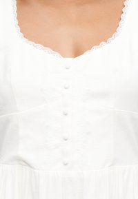 Λευκό στήθος φορέματος με διακόσμηση δαντέλας στη λαιμόκοψη, πέντε κουμπιά καλυμμένα με ύφασμα κατά μήκος του κέντρου και διακριτική κεντητή λεπτομέρεια με λουλούδια.