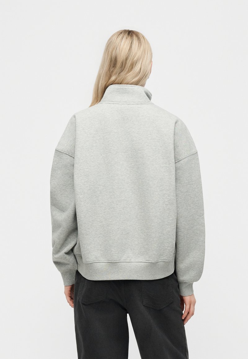 Persona con capelli lunghi e biondi che indossa una felpa grigia chiara con collo alto e pantaloni neri, in piedi di spalle contro uno sfondo semplice.