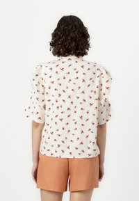 White blouse with small red floral print, puff sleeves, and a straight cut, paired with orange shorts. Textured fabric with subtle ridges.
