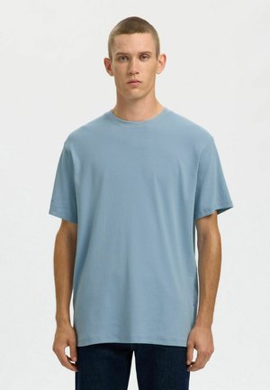 Jeune homme aux cheveux courts portant un t-shirt à manches courtes côtelé bleu clair uni et un pantalon foncé, debout devant un fond blanc.