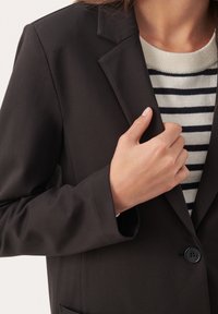 Černé sako s klasickými klopami, s jedním černým knoflíkem a hladkým povrchem. Pod ním pruhovaný svetr v bílé a námořnické modři.
