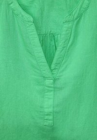 Grön linneblus med V-ringning, veckad detalj på baksidan och subtil sömnad längs kanterna. Mjuk textur över hela plagget.