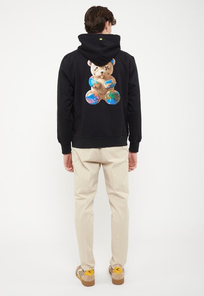 Άνδρας που φοράει μαύρο φούτερ με κουκούλα με γραφικό αρκουδάκι που κρατάει υδρόγειους στη πλάτη, μπεζ παντελόνι και μπεζ αθλητικά παπούτσια με κίτρινες λεπτομέρειες, στέκεται με την πλάτη στο βλέμμα.