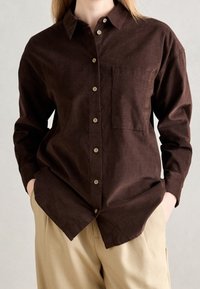 Camicia in velluto a coste marrone con bottoni sul davanti, colletto a punta, taschino sul petto e maniche lunghe, abbinata a pantaloni beige.