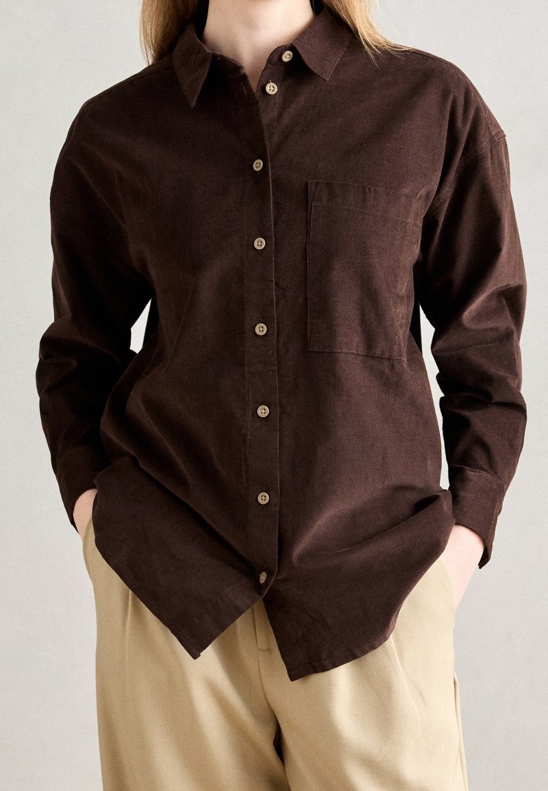 Camicia in velluto a coste marrone con bottoni sul davanti, colletto a punta, taschino sul petto e maniche lunghe, abbinata a pantaloni beige.