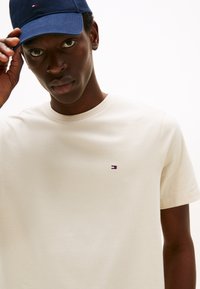 Kreemikas lühikeste varrukatega T-särk ümarate kaelusega ja vasakul rinnal väike tikitud logo punases, valges ja sinises värvitoonis.