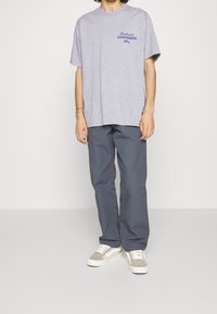 Person iført en lyslilla Carhartt Carrosserie Wip T-shirt, mørkegrå bukser og lysegrå og hvide sneakers, stående på et gråt gulv.