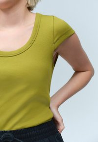 Maslinasto zelena rebrasta majica bez rukava s okruglim izrezom i kapicama, upotpunjena teksturiranim tamnim hlačama. Majica ima prilagođen oblik i glatku teksturu.