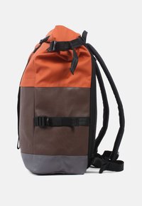 Rucksack mit orangefarbenem oberen Teil, braunem Körper und grauem Boden. Verfügt über einen Rollverschluss, verstellbare Träger und einen vorderen Schnallenverschluss.