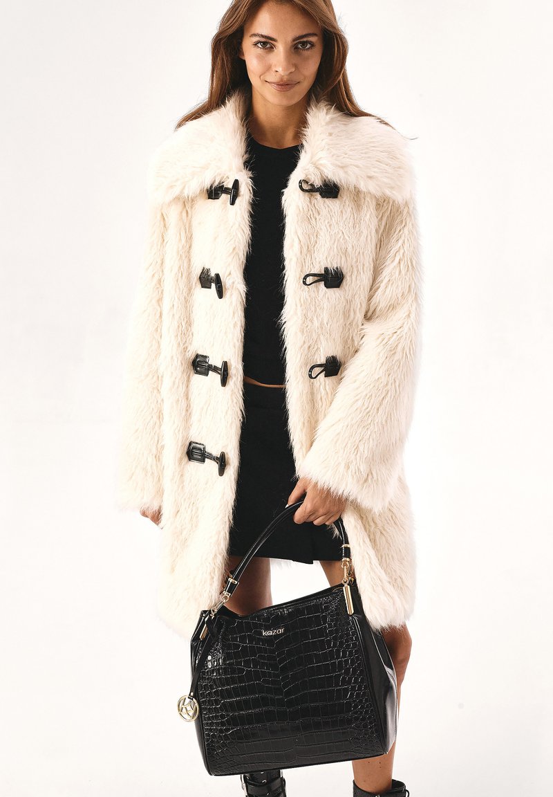 Cappotto in pelliccia sintetica color crema con ampio colletto, chiusure a toggle e maniche lunghe. Borsa a tracolla in pelle nera con motivo coccodrillo.