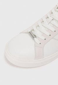 Zapatilla blanca con puntera y laterales de cuero liso, suaves acentos de ante gris, cordones blancos planos y una suela de goma texturizada.