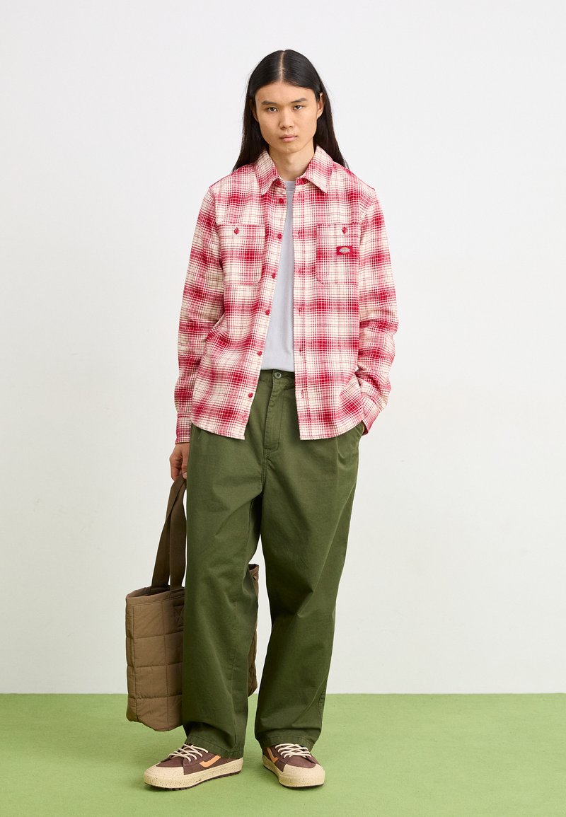 Czerwona i biała koszula w kratę flanelową, zielone spodnie o szerokich nogawkach, brązowe trampki z kremowymi podeszwami oraz brązowa pikowana torba na ramię.