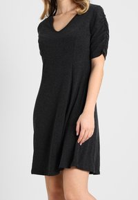 Zwarte, knie-lange jurk met een V-hals, korte gerimpelde mouwen, gemaakt van zachte stof. Heeft een losse, vloeiende silhouet.