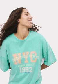 Ljusblå oversized T-shirt med ett bleknat orange tryck av "NYC", "Athletics Department" och "1992". Mjuk bomullstextur.