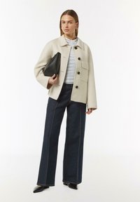 Cremfarbene Wollmischjacke mit einer Brusttasche, schwarzen Knöpfen, gestreiftem Oberteil, dunkelben weit geschnittenen Jeans und spitzen schwarzen Schuhen.