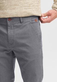 Pantalones de algodón grises con corte recto, con un botón marrón y un pequeño detalle de cuero cerca de la cintura. Textura suave, costuras sutiles.