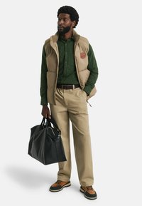 Tan puffer vest over donkergroen gespikkeld shirt, beige broeken, bruin en zwart schoenen, houdt een zwarte sporttas vast. Gladde, duurzame materialen.
