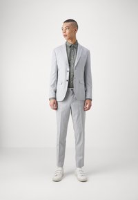 Traje gris claro con un diseño ajustado, dos botones y bolsillos de solapa, combinado con una camisa de botones verde oscuro y zapatillas blancas.